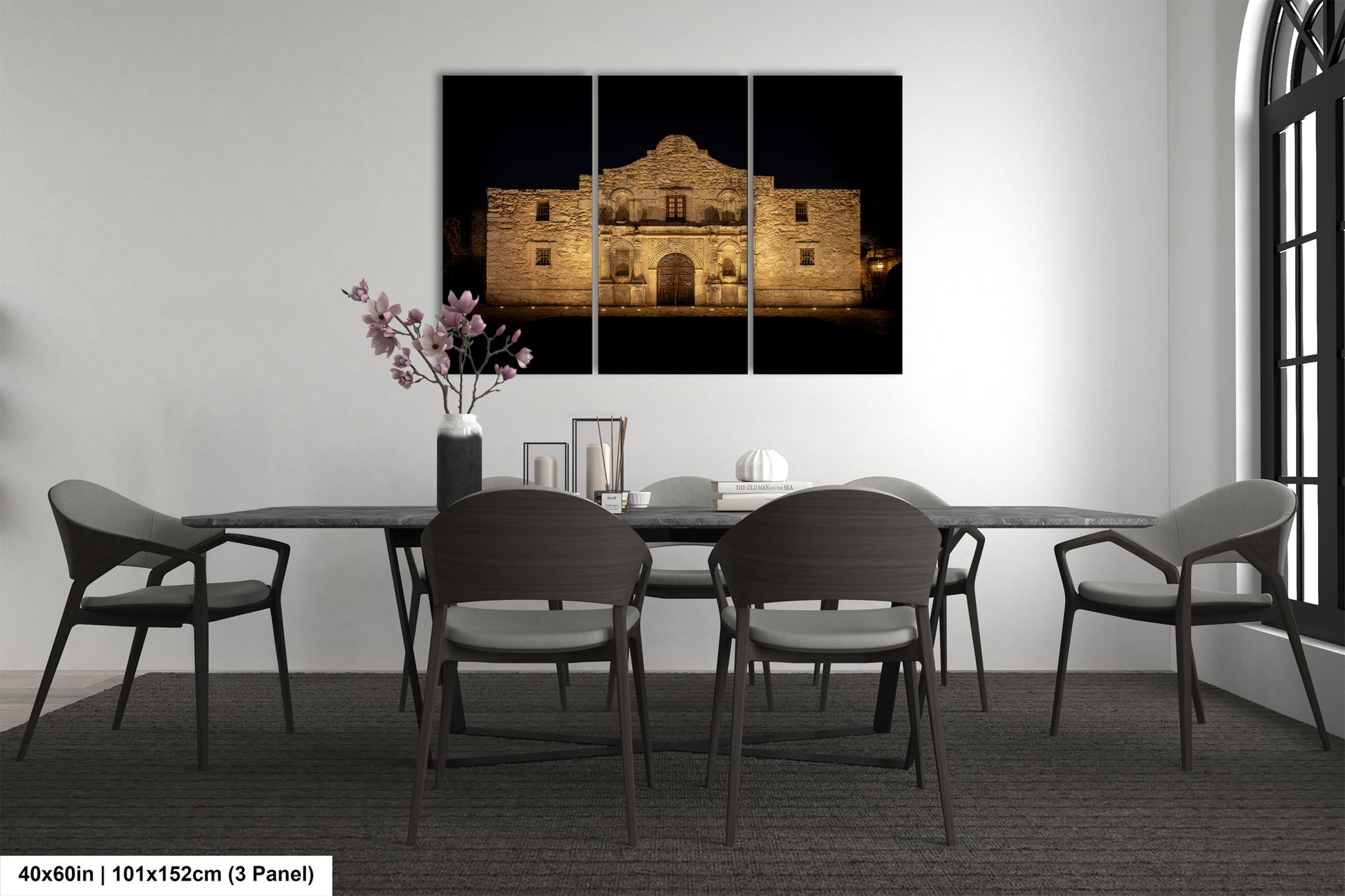 The image is a modern dining room with a large, illuminated photograph of the Alamo in the background. The room is furnished with a dining table and chairs, and there is a vase of flowers on the table.
