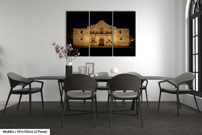 The image is a modern dining room with a large, illuminated photograph of the Alamo in the background. The room is furnished with a dining table and chairs, and there is a vase of flowers on the table.