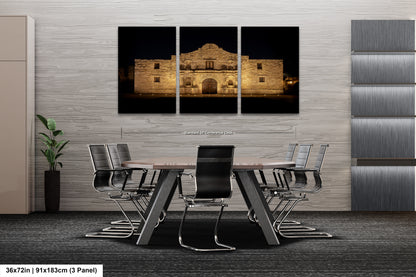 a modern office space with a large, illuminated photograph of the historic Alamo in the background. The photograph is divided into three panels, creating a collage-like effect. The office space includes a conference table surrounded by chairs, and a plant is visible in the corner.