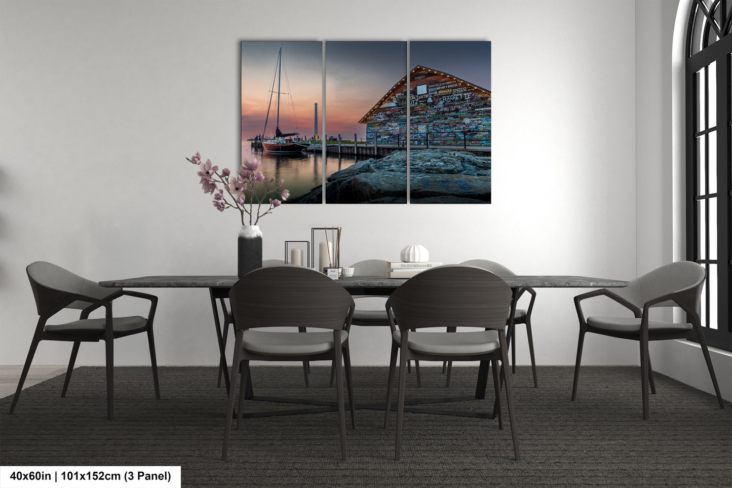 a modern dining room with a large, three-panel painting of a boat on a dock in the background. The painting is hung on the wall above a black dining table surrounded by six chairs. The room has a minimalist and contemporary design, with a vase of flowers and a book on the table adding a touch of color and personalization to the space.