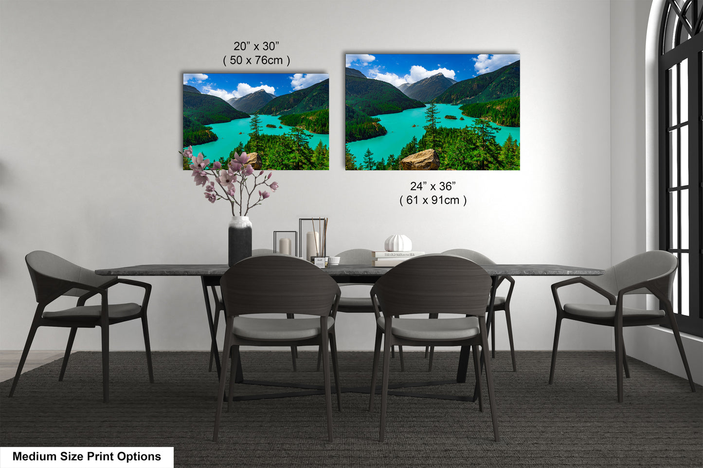 a dining room with a large black table surrounded by six chairs. Above the table, there are two framed landscape paintings depicting a scenic mountain view with turquoise water and green trees. The paintings are of different sizes, with one being larger than the other. The table and chairs are positioned in front of the paintings, creating a focal point in the room.