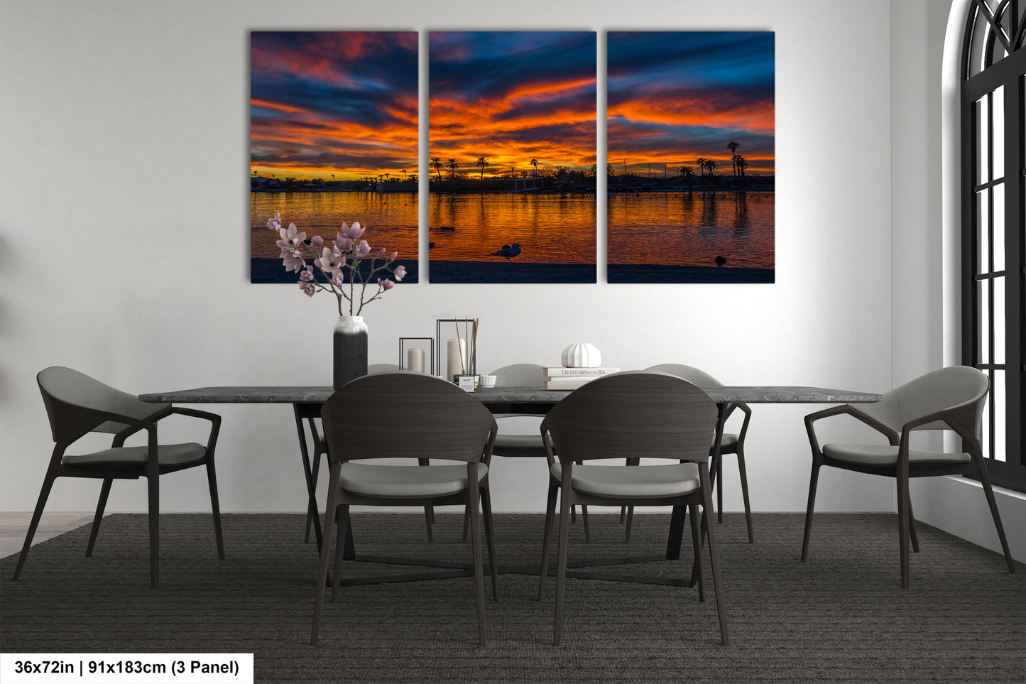 a modern dining room with a large, three-panel wall art depicting a serene sunset over a body of water. The art piece is framed in black and hangs above a black dining table surrounded by matching chairs. The table is set with a vase of flowers and a few decorative items, creating a cozy and inviting atmosphere.