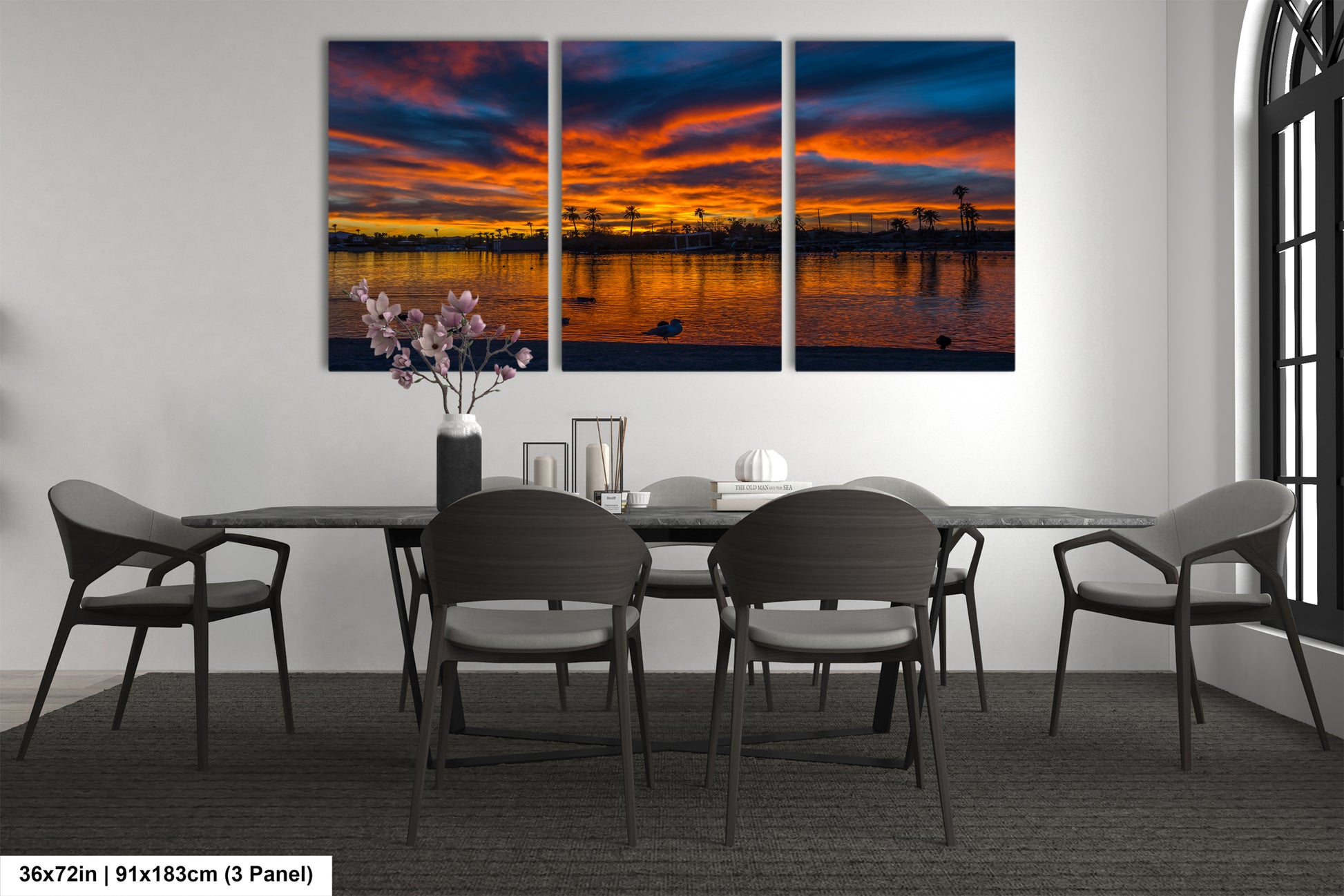 a modern dining room with a large, three-panel wall art depicting a serene sunset over a body of water. The art piece is framed in black and hangs above a black dining table surrounded by matching chairs. The table is set with a vase of flowers and a few decorative items, creating a cozy and inviting atmosphere.