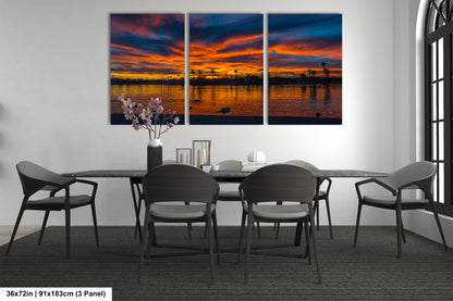 a modern dining room with a large, three-panel wall art depicting a serene sunset over a body of water. The art piece is framed in black and hangs above a black dining table surrounded by matching chairs. The table is set with a vase of flowers and a few decorative items, creating a cozy and inviting atmosphere.