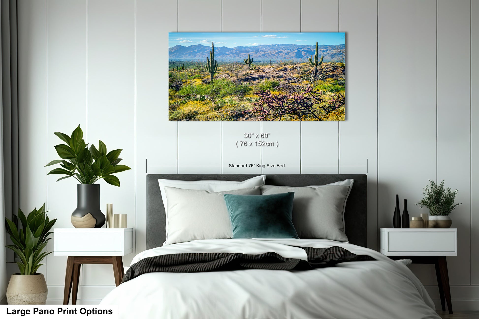 A large pano print depicting a desert landscape with cacti and mountains in the background, hanging on a wall above a bed with white bedding and pillows.