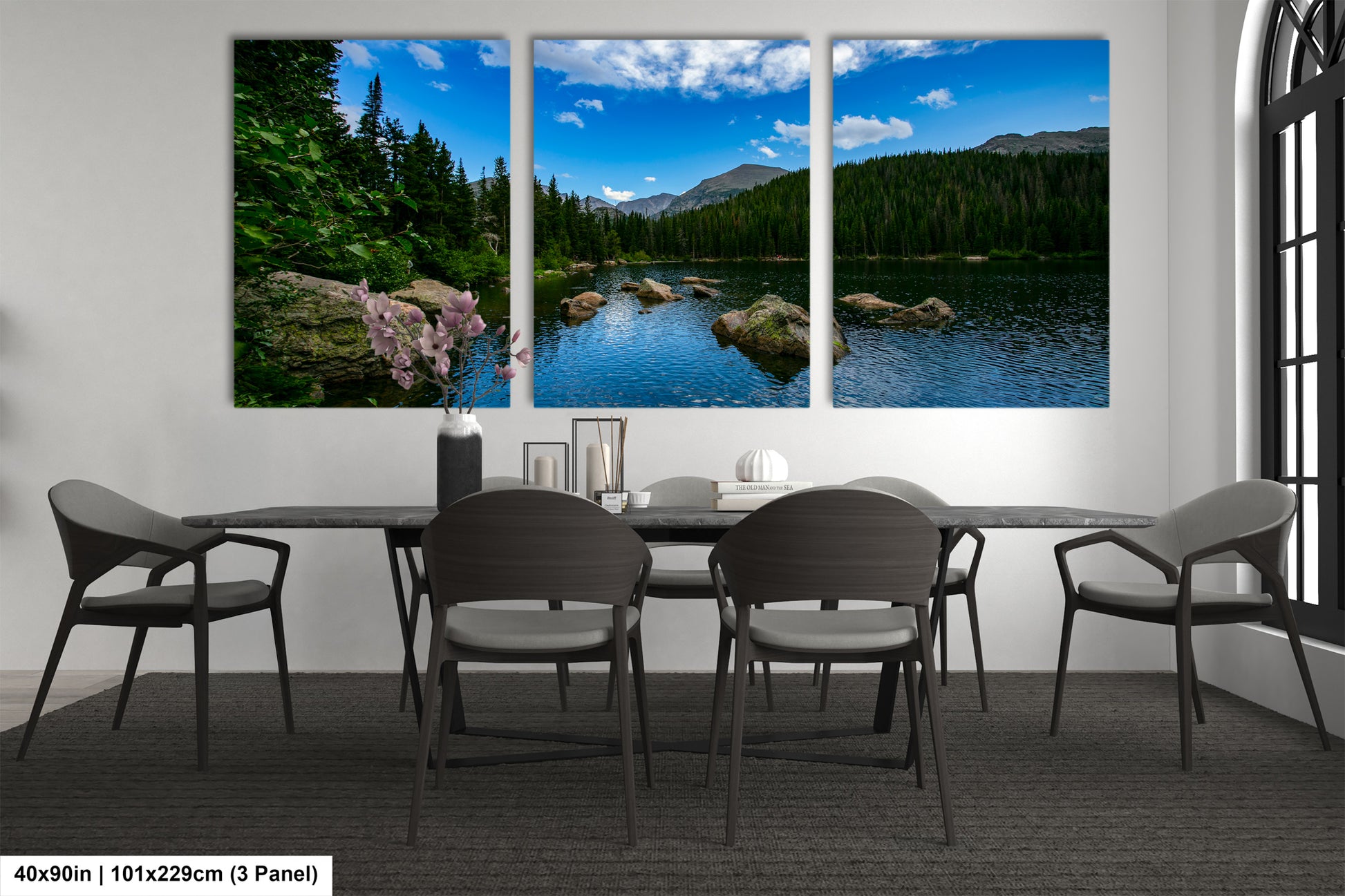 a modern dining room with a large window that offers a scenic view of a lake surrounded by mountains. The room is furnished with a black table and chairs, and there are decorative elements such as a vase and a book on the table.