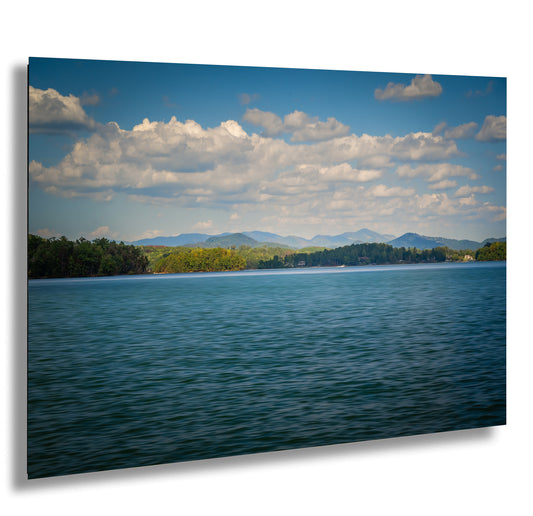 a serene lake surrounded by lush green trees and mountains, with a clear blue sky dotted with fluffy white clouds.