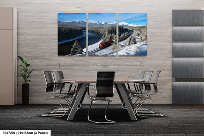A modern office space with a large, three-panel landscape painting depicting a snowy mountain scene with a train passing through it. The painting is mounted on a gray wall above a wooden table surrounded by chairs.