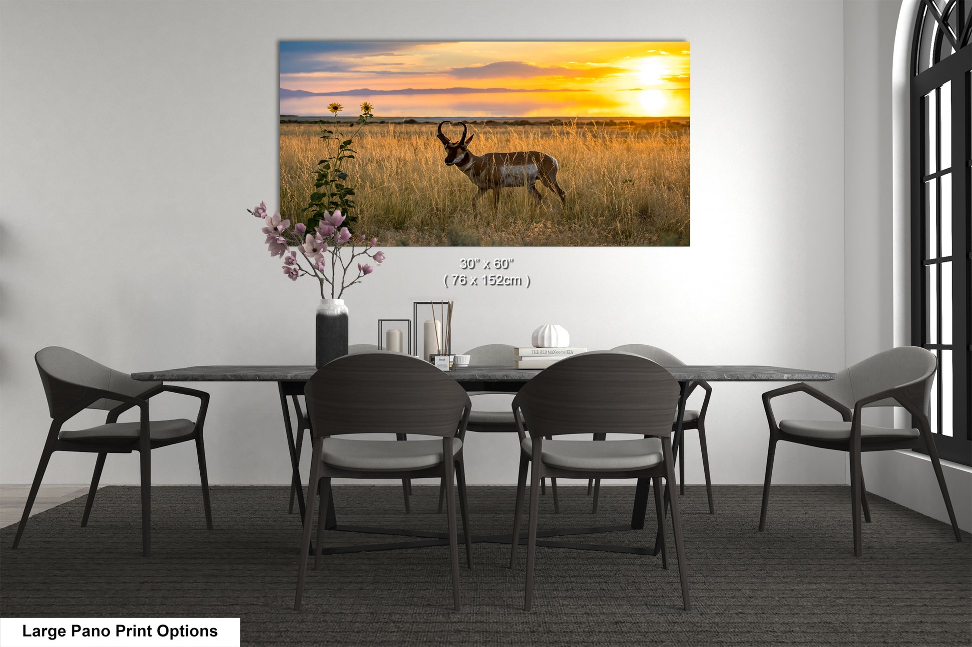 A large pano print depicting a wild animal in a grassy field at sunset, with a vase of flowers and a book on the table in the foreground.