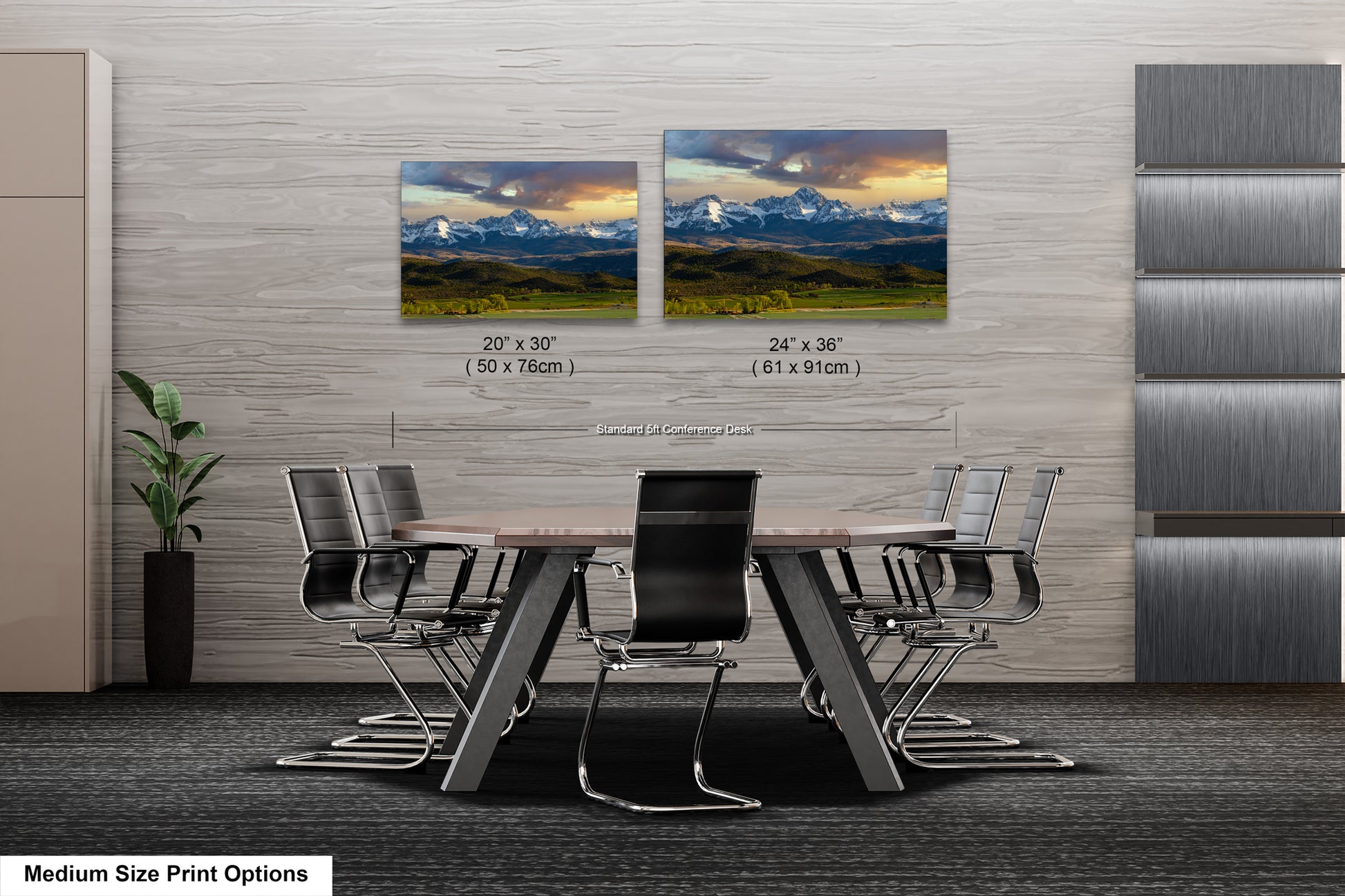 a modern office space with a large wooden table surrounded by black chairs. Two framed landscape paintings are displayed on the wall above the table, depicting a mountainous scene. The paintings are of medium size, with dimensions of 20 x 30" and 24 x 36" respectively. The office space also features a gray wall and a plant, creating a professional and comfortable atmosphere.
