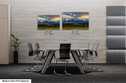 a modern office space with a large wooden table surrounded by black chairs. Two framed landscape paintings are displayed on the wall above the table, depicting a mountainous scene. The paintings are of medium size, with dimensions of 20 x 30" and 24 x 36" respectively. The office space also features a gray wall and a plant, creating a professional and comfortable atmosphere.