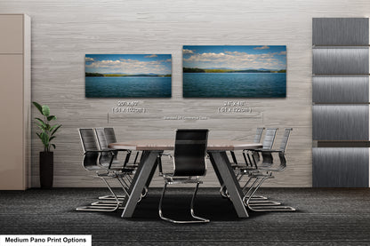 a modern office or conference room setting with a large wooden table surrounded by black chairs. Two framed landscape photographs are displayed on the wall above the table, depicting a serene body of water with a blue sky and clouds. The photographs are of similar size and framing, suggesting they are part of a collage or a single image. The overall design of the room appears to be minimalist and contemporary, with a focus on functionality and simplicity.