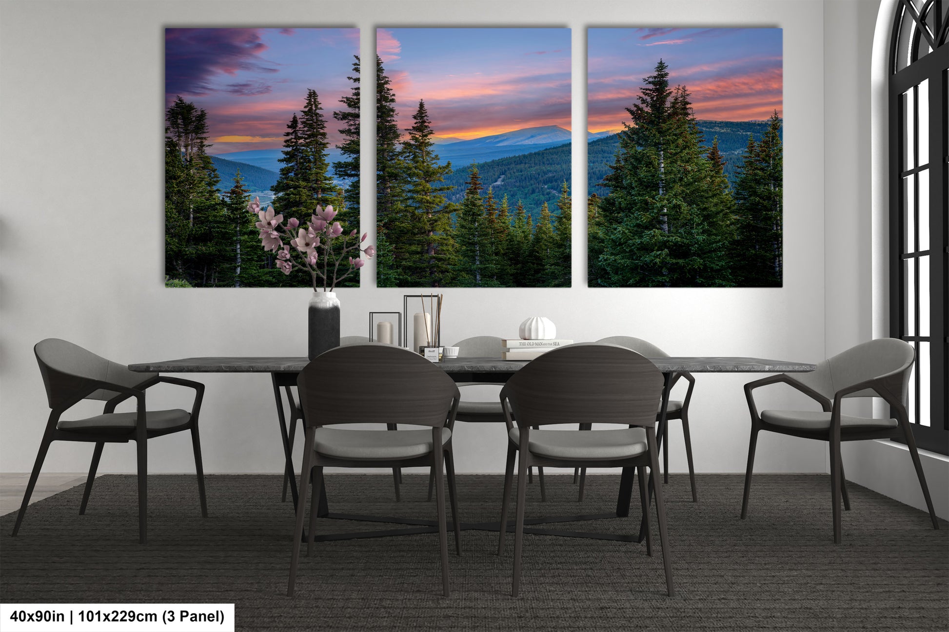 a modern dining room with a large window that offers a scenic view of a forest and mountains. The room is furnished with a black table and chairs, and there are three large paintings or photographs displayed on the wall, depicting a serene landscape with trees, mountains, and a colorful sky.