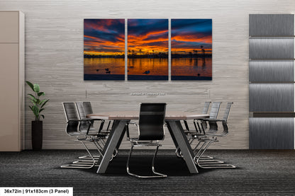 A modern office space with a large, three-panel landscape painting depicting a sunset over a body of water. The painting is mounted on a gray wall above a wooden table surrounded by chairs.