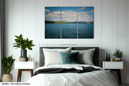 a bedroom with a large, high-quality, three-panel landscape painting hanging on the wall above the bed. The painting depicts a serene lake surrounded by mountains, creating a peaceful and picturesque scene. The bedroom has a comfortable bed with white linens and a gray headboard, and the room is decorated with potted plants and other decorative items, creating a cozy and inviting atmosphere.