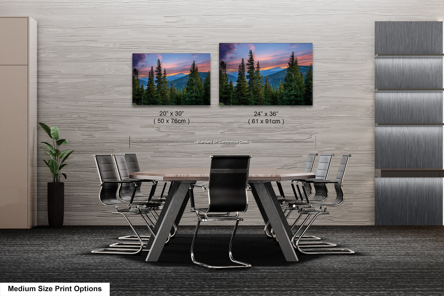 a modern office space with a large wooden table surrounded by black chairs. Two framed landscape paintings are displayed on the wall above the table, depicting a scenic mountain landscape with pine trees and a colorful sunset. The paintings are of medium size, with dimensions of 20 x 30" and 24 x 36" respectively.