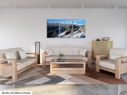 A modern living room with a large, three-panel landscape painting depicting a snowy mountain scene with a train passing through. The painting is hung on the wall above a wooden coffee table and matching chairs.