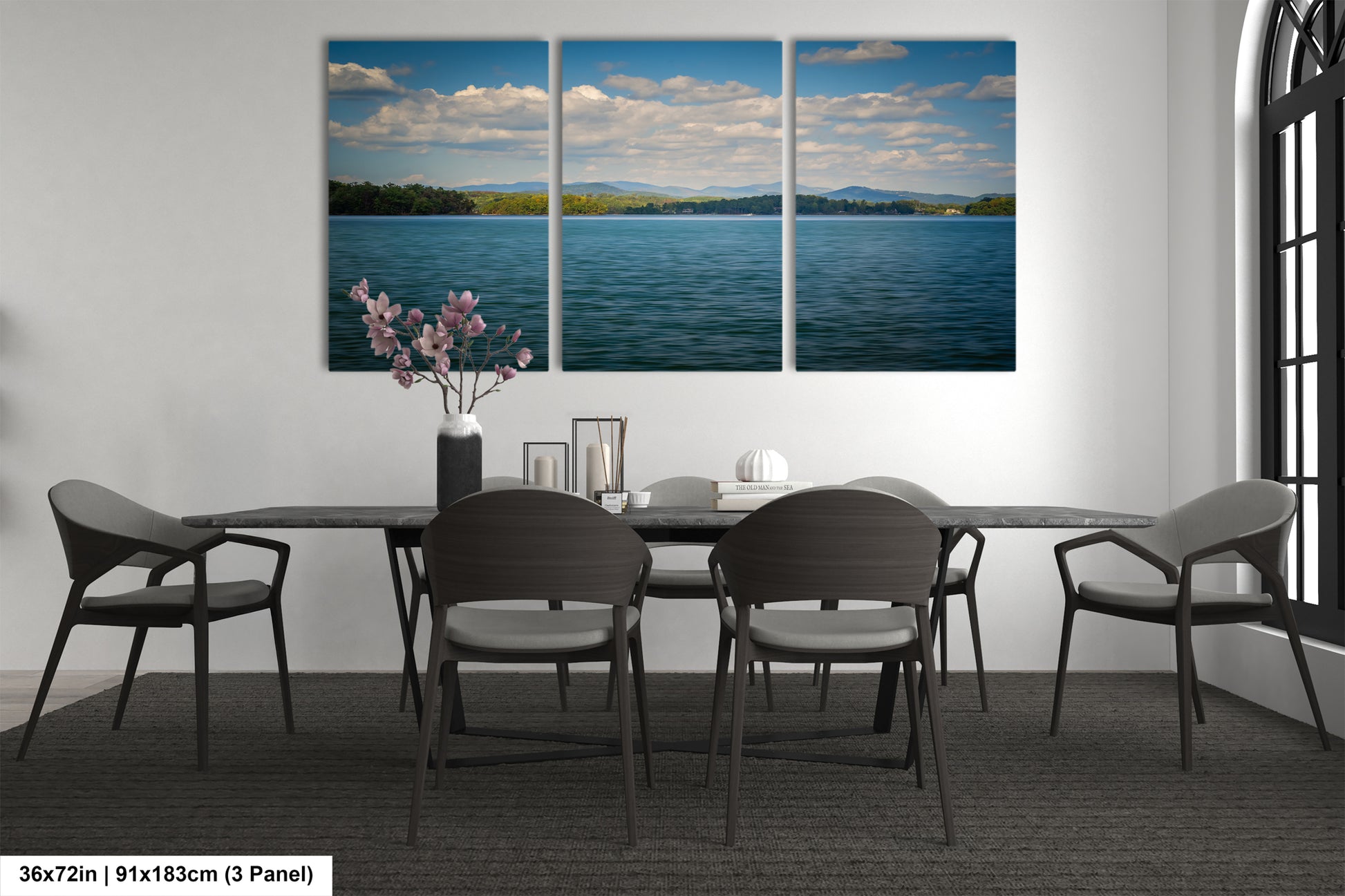 a modern dining room with a large wall-mounted painting depicting a serene lake scene with mountains in the background. The painting is divided into three panels, creating a sense of depth and perspective. The dining table is surrounded by six chairs, and there is a vase of flowers on the table, adding a touch of natural beauty to the space. The overall design and arrangement of the room create a harmonious and inviting atmosphere.