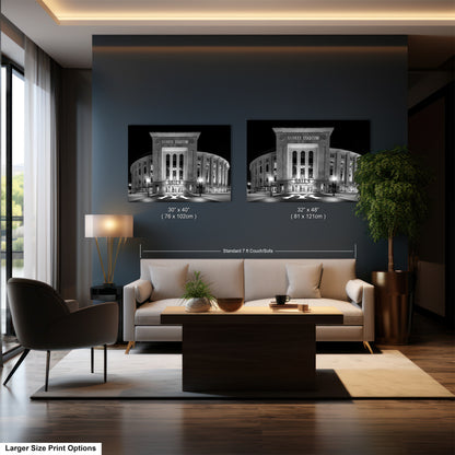 a modern living room with a large sofa, a coffee table, and a lamp. Above the sofa, there are two framed photographs of a stadium, which appear to be the Yankees stadium. The room has a dark color scheme, with a large window providing natural light.