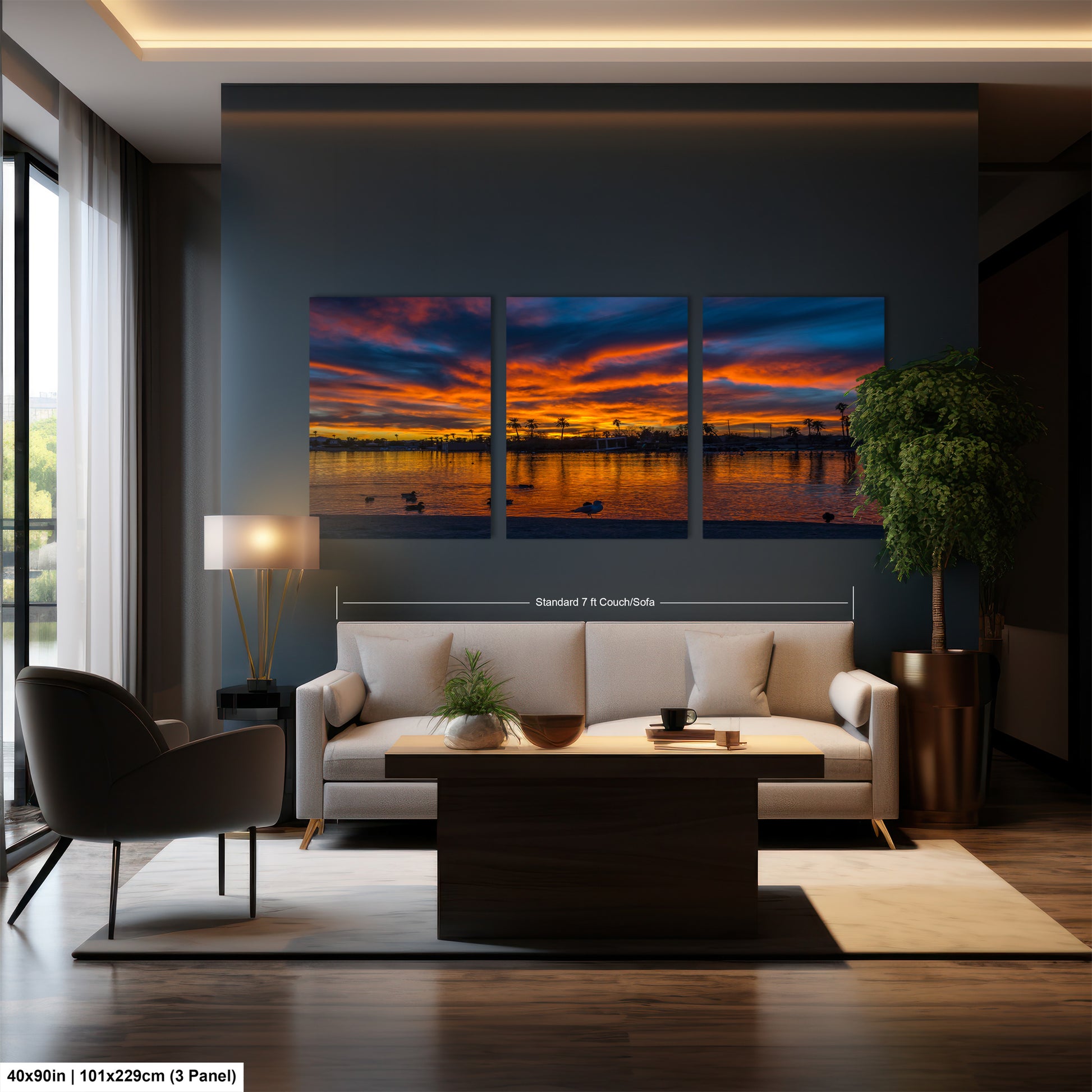 The image is a modern living room with a large, three-panel landscape painting depicting a sunset over a body of water. The painting is hung on the wall above a white sofa and a coffee table. The room also includes a chair, a lamp, and a potted plant, creating a cozy and inviting atmosphere.