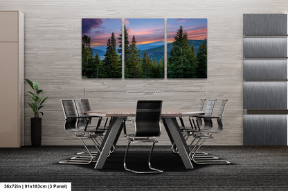 A modern office space with a large, three-panel landscape painting depicting a serene mountain scene with pine trees and a colorful sunset sky. The painting is mounted on a gray wall above a wooden table surrounded by black chairs.