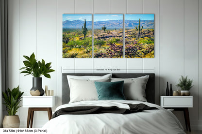 A bedroom with a large bed, nightstands, and potted plants. Above the bed is a large canvas print depicting a desert landscape with cacti and mountains in the background.