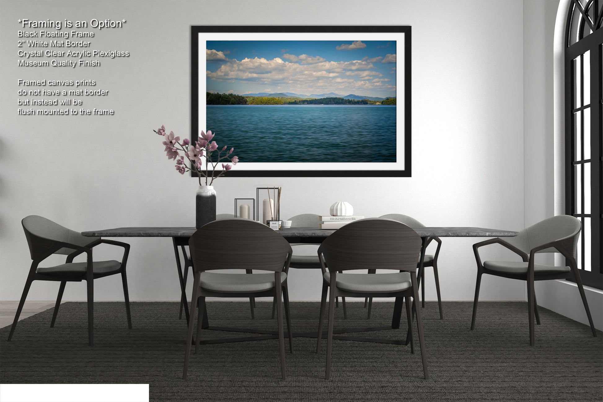 a modern dining room with a large black dining table surrounded by six matching chairs. Above the table, there is a framed picture of a serene lake with a cloudy sky. The room has a minimalist and elegant design, with a vase of flowers on the table and a bookshelf in the background.