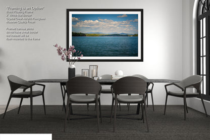 a modern dining room with a large black dining table surrounded by six matching chairs. Above the table, there is a framed picture of a serene lake with a cloudy sky. The room has a minimalist and elegant design, with a vase of flowers on the table and a bookshelf in the background.