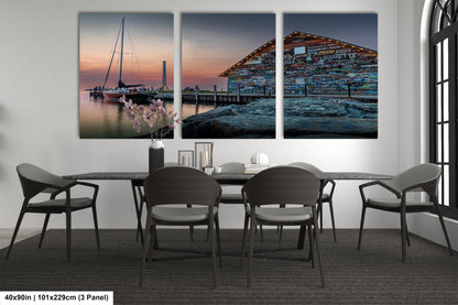 The image is a triptych of a seascape with a sailboat and a building with a colorful facade, displayed on a wall above a bed with white linens and a gray headboard.